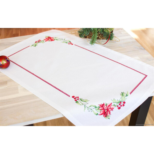 A rectangular table runner with a white background is intricately embroidered in cross stitch with red poinsettias and green leaves on the corners and edges. The embroidery pack from Luca-s lies on a light-colored wooden table, decorated with a fir branch and a red ornament on one side.