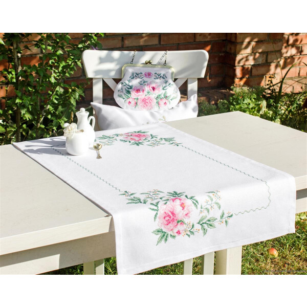 Una mesa blanca con un camino de mesa bordado con flores, rosas rosas y hojas verdes en un jardín. Sobre la mesa hay una tetera blanca, un pequeño jarrón con flores blancas y una copa plateada. Un paquete de bordados de Luca-s a juego con el cojín floral de la silla de al lado completa el encantador conjunto.