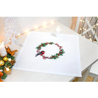 A white placemat from a Luca-s embroidery pack shows an embroidered wreath of red berries and green foliage with a small bird, all carefully worked in cross-stitch. The table is adorned with candles and festive decorations, including a white ornamental tree and a small bundle of golden baubles, pine cones and orange ribbon.