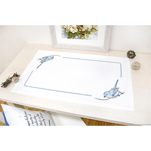 Een witte tafel is gedecoreerd met een geborduurde tafelloper, waarop twee blauwe vogels zitten op takken in tegenovergestelde hoeken. Aan de achterkant van de tafel hangt een ingelijst bloemenschilderij en eromheen zijn kleine decoratieve voorwerpen geplaatst. Op de voorgrond is een blauw-wit kussen te zien dat deel uitmaakt van een borduurpakket van Luca-s.