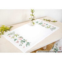 A white rectangular table runner on a light-colored wooden table with green leaves and pink floral embroidery on the edges. In the center of the runner is a small bouquet of flowers. In the background are more flowers and a chair with similar embroidery from Luca-s embroidery pack.