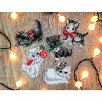 Five cat ornaments decorated with cross-stitch lie on a wooden surface, surrounded by glowing fairy lights. The cats have different colors - black and white, tabby and white with colorful bows and Christmas decorations. Each cat, carefully crafted from a Letistitch embroidery pack, is decorated with festive accessories such as holly and ornaments.