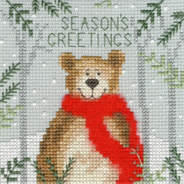 A cross stitch embroidery pack from Bothy Threads featuring a smiling bear wearing a bright red scarf. Fir branches frame the bear on both sides. Snowflakes adorn the gray background and the text Seasons Greetings is embroidered above the bears head.