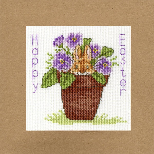 Cross stitch image on brown paper showing a bunny peeking out of a pot of purple flowers. Happy Easter is stitched around the Bothy Threads embroidery pack, with Happy Easter on the left and Easter on the right. The pot is standing on green grass with leaves and flowers sticking out of it.