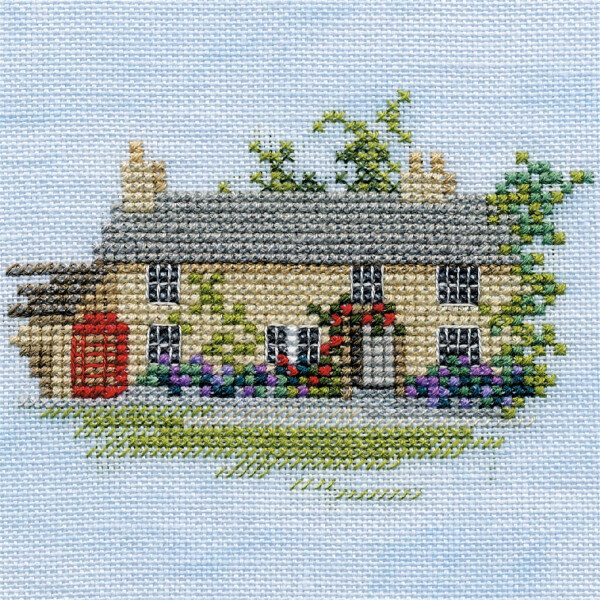 Cross stitch picture of a picturesque cottage with a gray roof, beige walls and several chimneys. This charming embroidery pack from Bothy Threads is decorated with climbing plants and surrounded by colorful flowers. To the left of the cottage is a red telephone box against a light blue background.