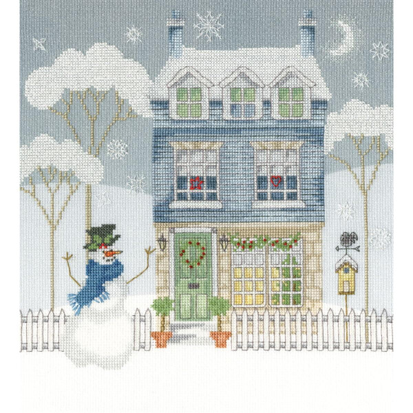 Eine im Kreuzstich genähte Winterlandschaft (Stickpackung, Bothy Threads) zeigt ein hellblaues dreistöckiges Haus, das mit einem Weihnachtskranz geschmückt ist und von kahlen Bäumen und Schneeflocken umgeben ist. Vor dem Haus ist ein weißer Lattenzaun, ein Schneemann mit Mütze, Schal und Karottennase. Auf der rechten Seite thront ein Vogelhäuschen.