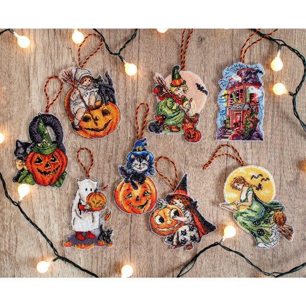 Die Halloween-Ornamente der Letistitch Stickpackung sind auf einer Holzoberfläche angeordnet und von leuchtenden Lichterketten umgeben. Zu den Ornamenten gehören Kürbisse, Hexen, ein Spukhaus, ein Geist, der einen Kürbis hält, eine Krähe auf einer Kürbislaterne und eine Mondsichel mit einer Fee.