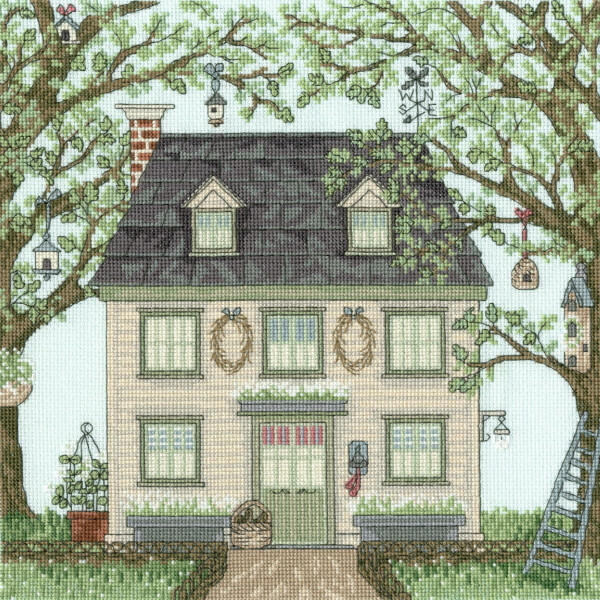 Dies ist eine Bothy Threads Stickpackung eines malerischen zweistöckigen Hauses mit grauem Dach und grünen Akzenten. Um das Haus herum stehen Bäume mit Vogelhäuschen und an einem lehnt eine Leiter. Das Haus hat einen gemauerten Schornstein, Kränze und Topfpflanzen, die seinen Charme noch verstärken. Ein Gehweg führt zur Haustür.