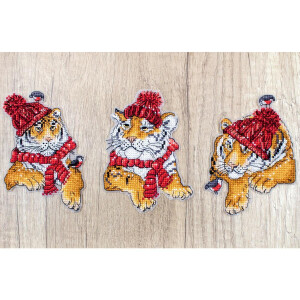 Three cartoon-style tiger cubs wear red winter hats and scarves with little birds on the tassels. The cubs, depicted in cross stitch on a wooden background, are seated and appear cozy, each has a unique pose and expression and features playful and festive details. This scene can be seen in the Letistitch embroidery kit.
