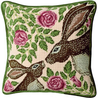 An embroidered picture of two brown rabbits with pink inner ears and detailed eyes facing each other on a light pink background is embroidered on a square cushion. Surrounding them are green leaves and pink roses. The Bothy Threads embroidery pack has green piping around the edges, showing intricate cross stitch craftsmanship.