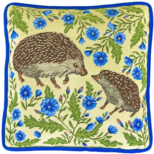 A decorative cushion with an embroidery pack design from Bothy Threads shows two hedgehogs surrounded by blue flowers and green leaves on a beige background. The cushion has a blue border. The larger hedgehog is facing left, while the smaller one is facing the larger one, creating a charming nature-inspired scene.