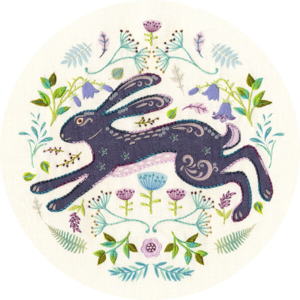Un lapin bleu foncé aux motifs compliqués, brodé de fils lumineux, bondit entre une rangée de fleurs et de vrilles colorées sur un fond de tissu crème. Cette scène du kit de broderie Bothy Threads est pleine de nuances de violet, de vert et de bleu qui contribuent à un design bizarre et détaillé.