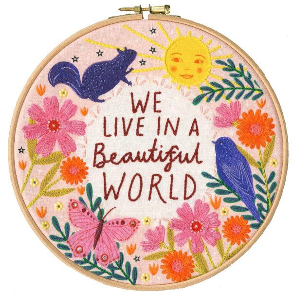 Kleurrijk borduurwerk in een houten frame met de tekst WE LIVE IN A BEAUTIFUL WORLD omringd door een zon, rode en roze bloemen, groene bladeren, een paarse eekhoorn, een roze vlinder en een blauwe vogel op een crèmekleurige stoffen achtergrond, perfect voor iedereen die van een gedetailleerd borduurpakket van Bothy Threads houdt.