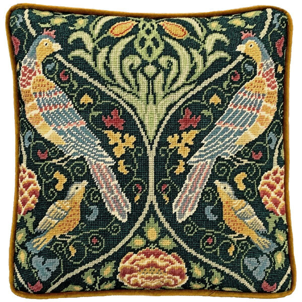 A square cushion with intricate needlepoint embroidery, similar to a Bothy Threads embroidery pack, features two mirrored birds with colorful plumage against a dark green background. The design is embellished with floral and leaf patterns in shades of green, yellow, red and blue. The cushion has a yellow piped edge.