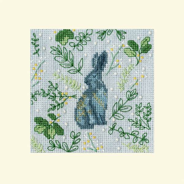 Stickerei einer Kaninchensilhouette in marineblauem und grauem Kreuzstich, umgeben von grünen Blattzweigen mit Akzenten aus gelben Blumen auf hellblauem Stoffhintergrund. Die Stickpackung von Bothy Threads ist in einem quadratischen Rahmen eingeschlossen.