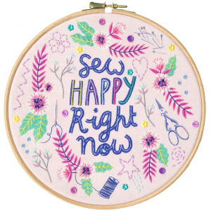 A decorative embroidery hoop with colorful stitching features the phrase Sew Happy Right Now in the center. Surrounding the text are various embroidered designs, including flowers, leaves, sewing needles, scissors, a spool of thread and buttons - all reminiscent of a vibrant Bothy Threads embroidery pack on a bright fabric background.