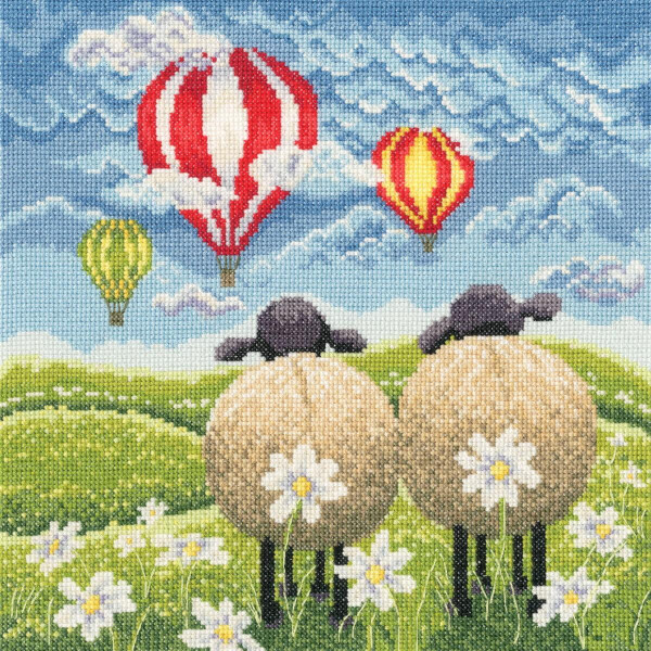 Two sheep with black heads and woolly bodies stand with their faces turned away in a meadow strewn with white daisies. The sky is clear with a few clouds and shows four colorful hot air balloons in red, yellow and green floating over rolling green hills. The scene is bright and whimsical, perfect for a Bothy Threads embroidery pack.