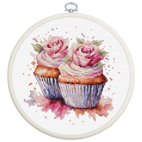 A charming cross stitch artwork from an embroidery kit by Luca-s, framed in a round frame, features two cupcakes with intricate pink rose frosting. The cupcakes are adorned with green leaves on the roses and are set against a white fabric background with scattered pink and purple stitch patterns around them.
