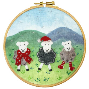 An embroidery frame shows three sheep in a grassy landscape with green hills and a blue sky. One sheep is wearing a red scarf, another a Santa hat and black sweater, and the third has a gray sweater and striped socks. The detailed stitching in this embroidery pack from Bothy Threads adds texture to the whimsical scene.