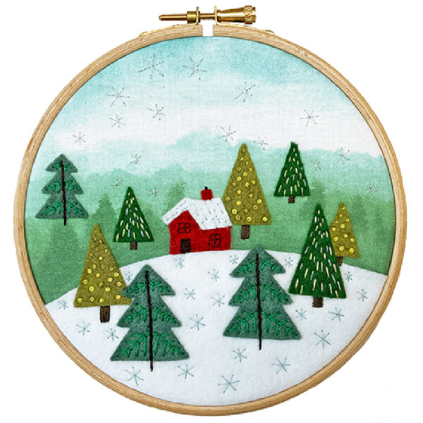 Ein runder Stickrahmen zeigt eine Winterlandschaft mit einer roten Hütte, umgeben von grünen Filzkiefern in einer verschneiten Landschaft. Im Hintergrund fällt ein hellblauer Himmel mit weißen Schneeflocken. Der Stoff ist straff gespannt und gibt die verschneite Umgebung der Bothy Threads Stickpackung wieder.