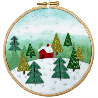 A round embroidery hoop shows a winter landscape with a red hut surrounded by green felt pines in a snowy landscape. A light blue sky with white snowflakes falls in the background. The fabric is tightly stretched and reflects the snowy surroundings of the Bothy Threads embroidery pack.