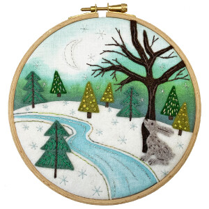 Embroidery depicting a snowy landscape with a crescent moon. Shows a flowing river, evergreen trees and a leafless tree. A gray rabbit sits next to the tree. The scene, framed in a wooden embroidery hoop, has a background of green and white with decorative snowflakes - an exquisite creation from Stickpackung by Bothy Threads.