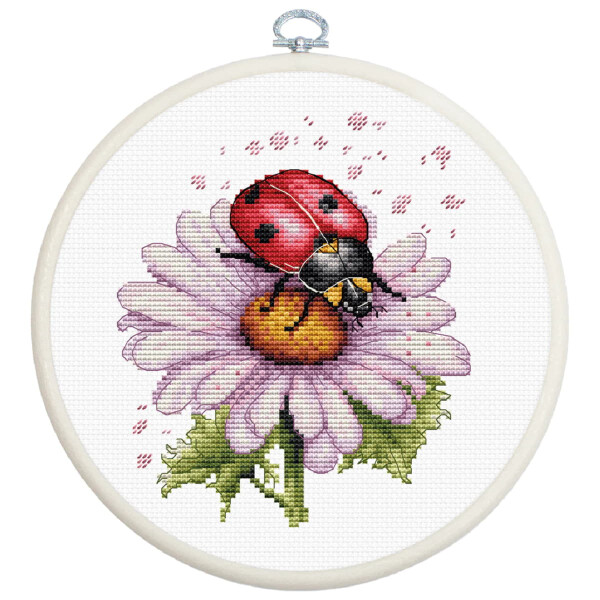A detailed picture of Luca-s embroidery pack with a ladybug on a pink daisy, framed in a white embroidery hoop. The ladybug is red with black dots and the daisy has purple petals with a yellow center. Green leaves are visible under the flower. Small pink stitches surround the ladybug and suggest movement.