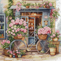 Een vintage fiets met gevlochten manden vol roze bloemen staat voor een charmant winkeltje. De winkel, die Lucas borduurpakketten verkoopt, is versierd met nog meer bloeiende bloemen in potten en hangplanten. De deur staat open en onthult een gezellig interieur met planken. Het tafereel is vol groen en kleurrijke bloemen.