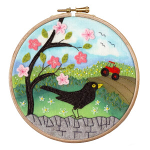An embroidery hoop shows a lively landscape scene with a black bird standing on a stone path in the foreground. Behind it, a tree blooms with pink flowers and green leaves. In the background, a red tractor drives on a dirt road under a bright blue sky with birds in flight. This charming embroidery pack from Bothy Threads features intricate details that are perfect for any cross stitch enthusiast.