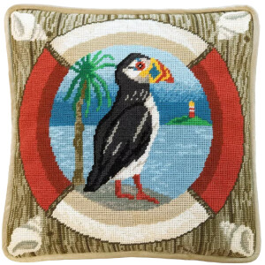 A square cushion with a colorful needlepoint design of a puffin standing next to a palm tree and water, in a round frame reminiscent of a lifebuoy. The Bothy Threads embroidery pack features an island with a lighthouse in the background and has a beige border with intricate corner details.