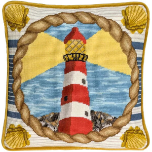 A square cushion with needlepoint embroidery, made using the cross-stitch technique, shows a red and white striped lighthouse emitting yellow rays of light. The lighthouse stands on rocky terrain against a blue sky with white clouds. The border consists of shells and a rope pattern, all outlined in yellow. This exquisite piece is available as an embroidery pack from Bothy Threads.