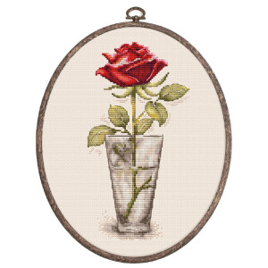 A piece of embroidery in an oval wooden frame shows a single red rose in a glass. The intricate embroidery features detailed shading on the petals, leaves, stem and glass, creating a realistic look. This Luca-s embroidery pack uses a light, neutral fabric background to highlight the vibrant colors of the rose.