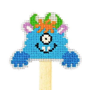 An embroidery pack design by Bothy Threads on a rectangular canvas features a playful blue monster with a single eye, green horns, orange hair and purple ears, showing a wide grin with prominent teeth. The embroidered image is attached to a wooden stick, which resembles a doll, against a plain white background.