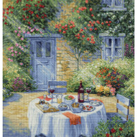 A picturesque scene with an outdoor table set for a meal, set in a lush garden with bright flowers. The table, covered with a white cloth decorated with Lucas embroidery packet embroidery, has room for plates, cups, a wine bottle, glasses and various food items. A brick wall with hanging rose bushes and a blue door can be seen in the background.