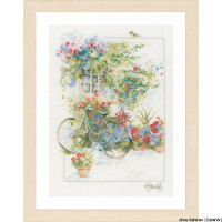 A framed artwork by Lanarte Stickpackung depicts a picturesque scene with an old bicycle parked in front of a building. The bicycle has a basket full of colorful flowers and potted plants with bright blooms surround the scene. The light-colored wooden frame complements it, while the artists signature can be seen at the bottom right.